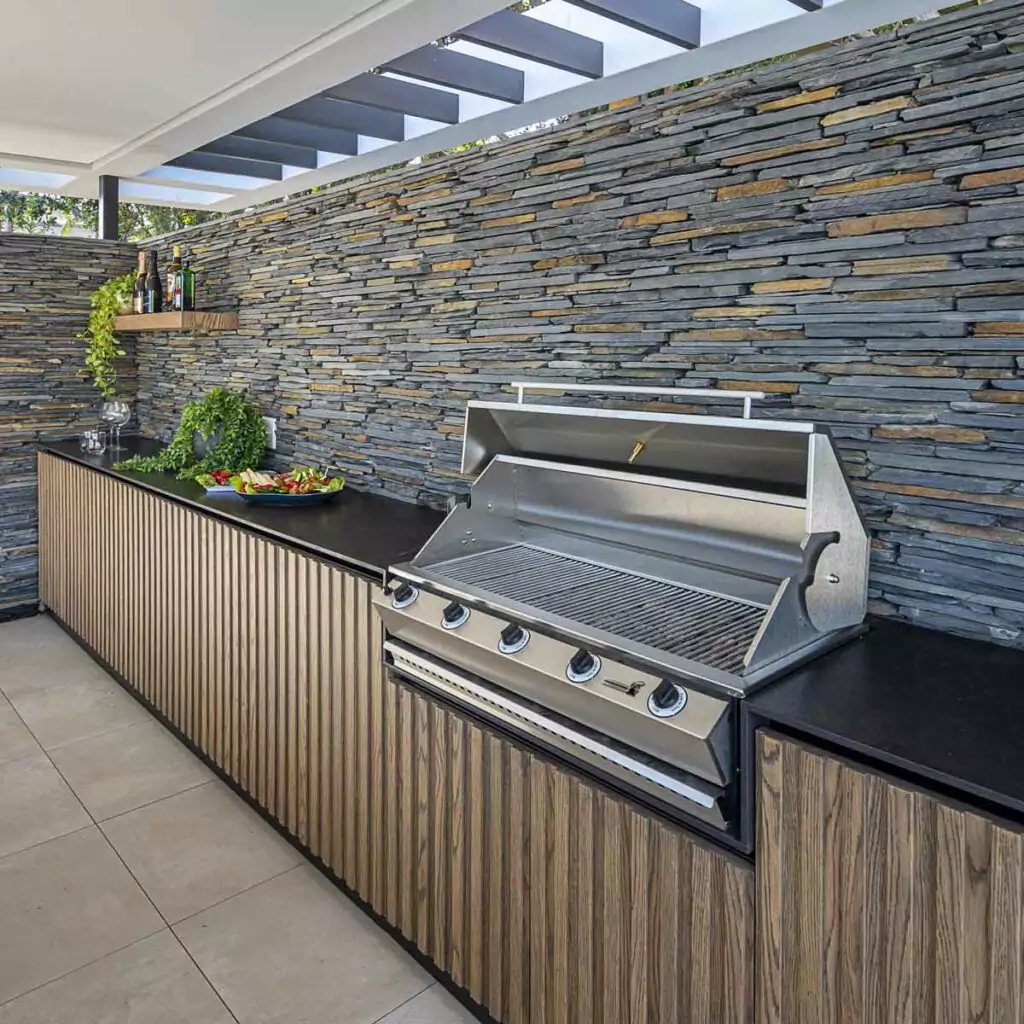 Outdoor kitchen with a stainless gas grill, stone accent wall, and wooden cabinets; plate of appetizers on the counter.